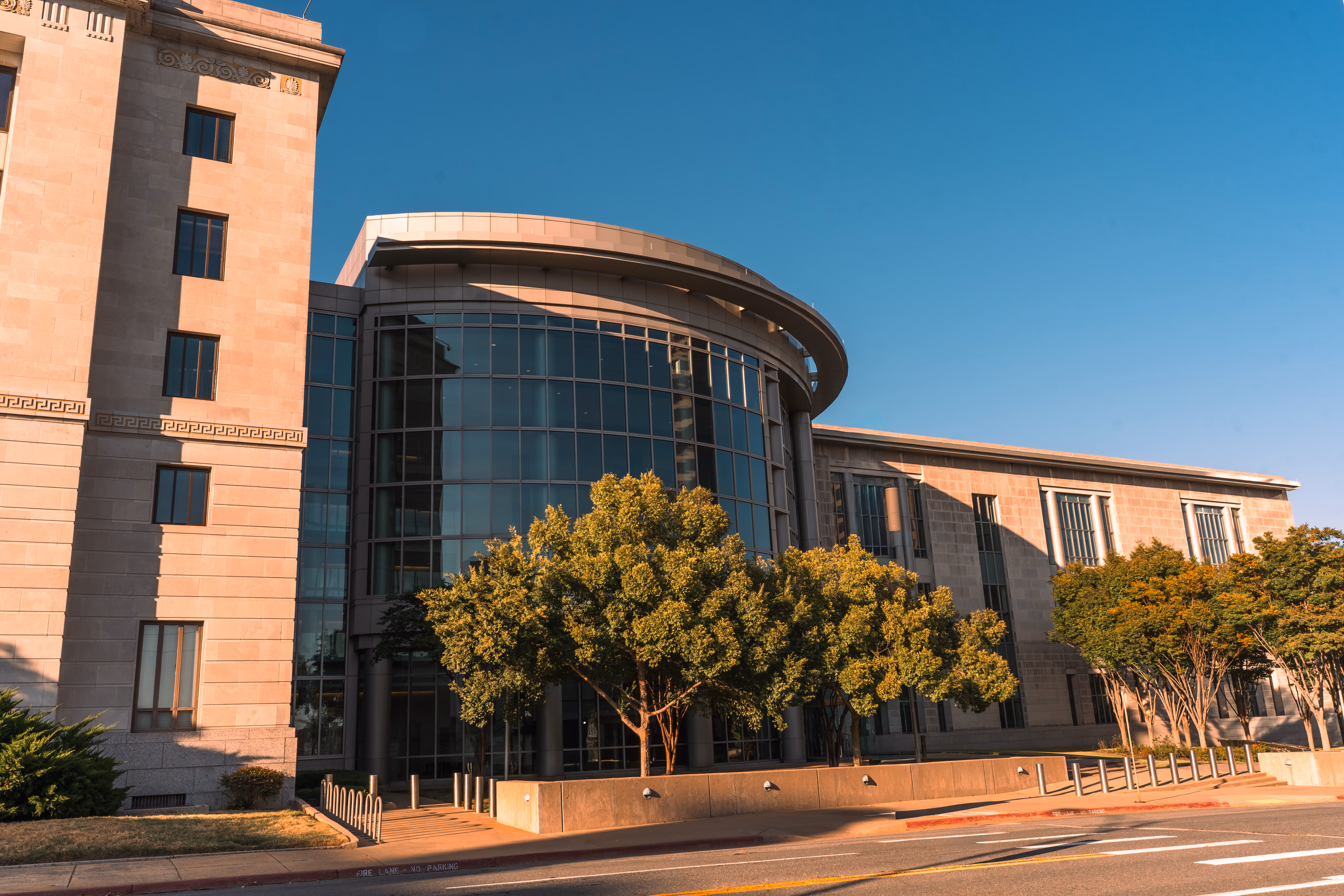 A glazed courthouse rotunda beside its limestone wing, screened by street trees