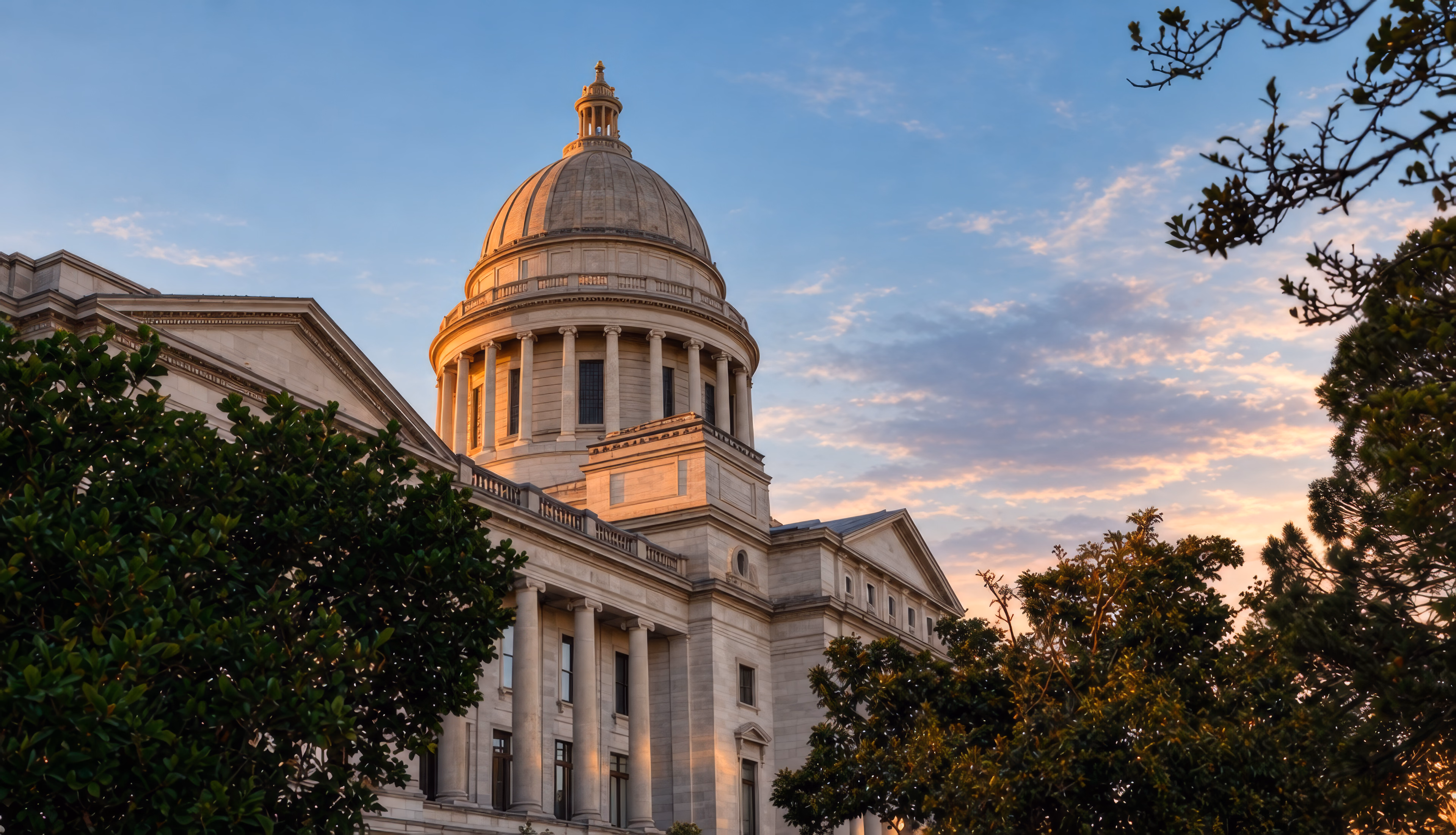 The dome of the Arkansas State Capitol at sunset, framed by trees